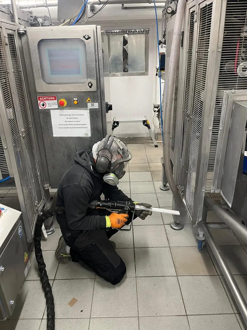 Industriereinigung in der Lebensmittelindustrie: Trockeneisstrahlen im unteren Bereich einer Produktionsmaschine mit Vollmaske, Gehörschutz und Handschuhen